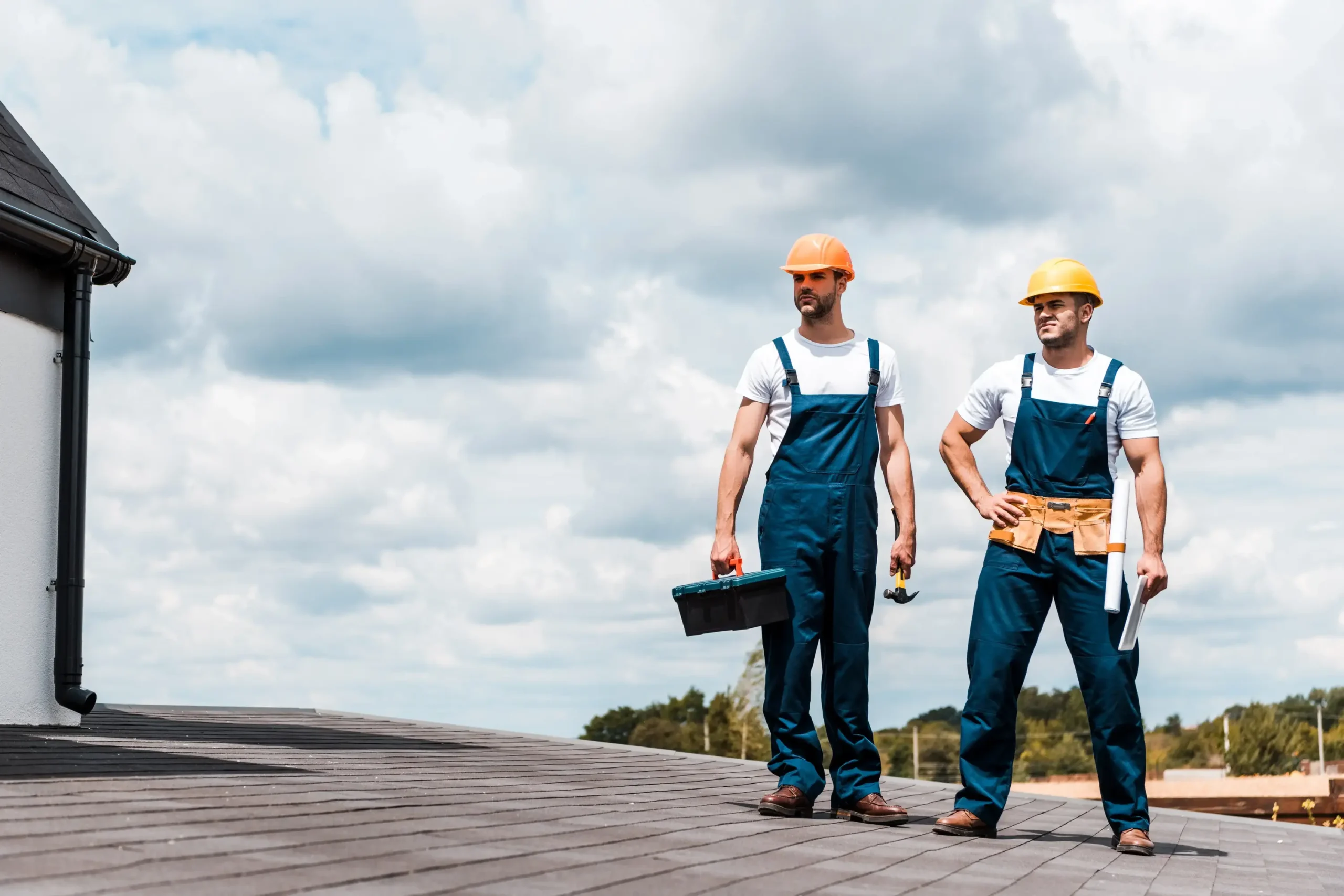 Two construction workers in hard hats and overalls stand on a rooftop, holding tools and blueprints.
